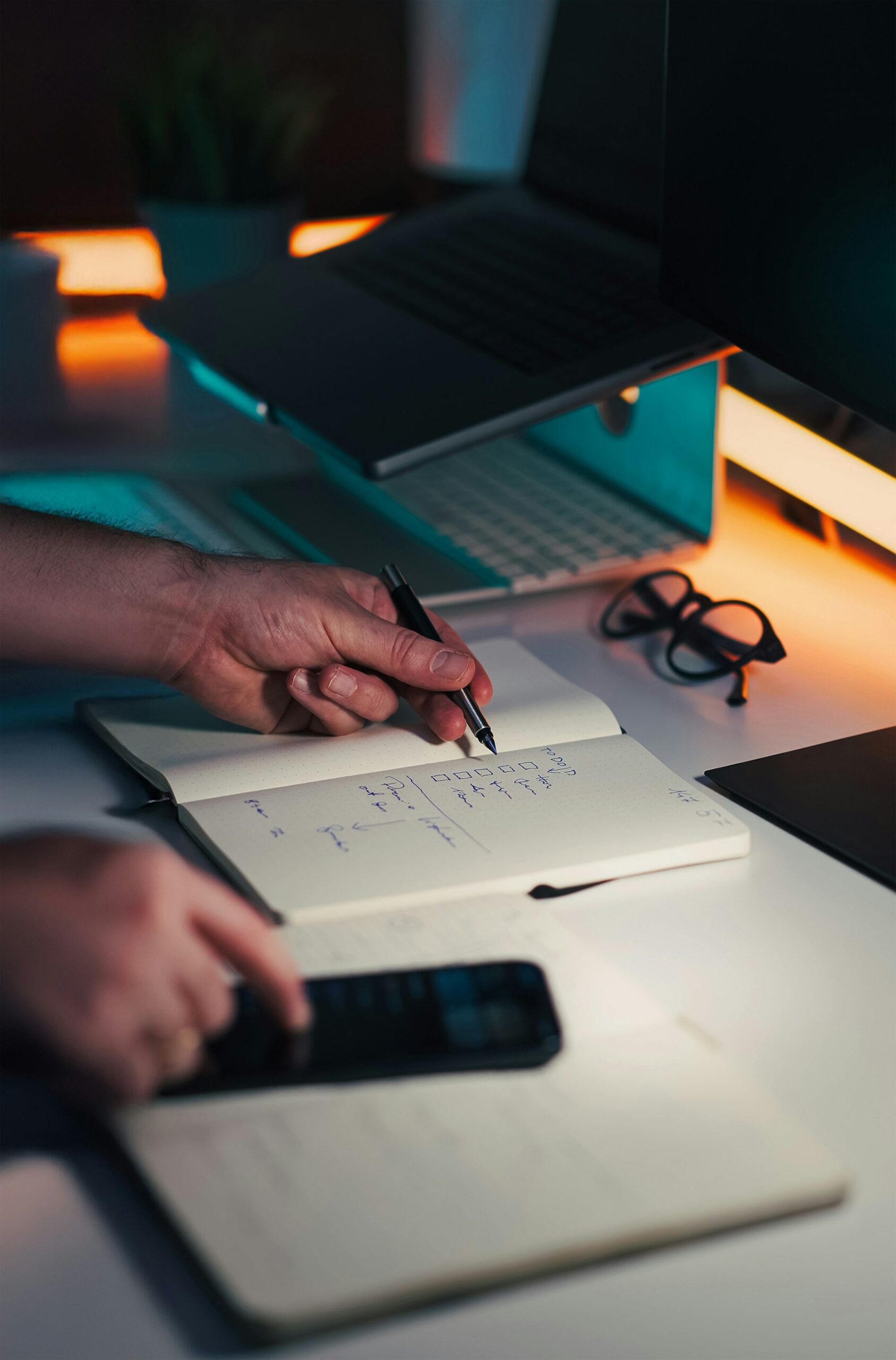 Hands creating a checklist beside a laptop and phone, representing workflow improvement and automation.